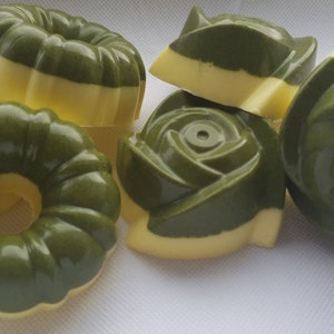 Può includere: Un assortimento di saponi fatti a mano in varie forme, tra cui disegni floreali e a ciambella. I saponi sono bicolore, con una parte superiore verde scuro e una base giallo chiaro. I saponi sono disposti su una superficie bianca.