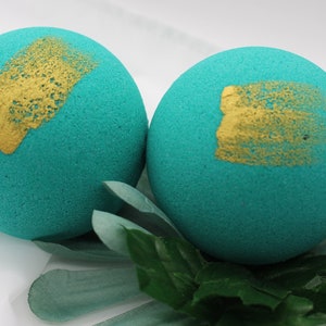 Puede incluir: Dos bombas de baño turquesas con trazos de pintura dorada, sobre hojas verdes y una superficie blanca. Las bombas esféricas tienen una superficie texturizada y un acento dorado decorativo. Primer plano.