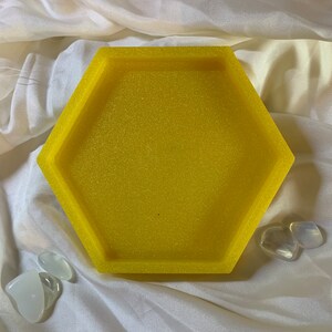 Puede incluir: Una bandeja hexagonal amarilla con un acabado brillante. La bandeja está vacía y se encuentra sobre una superficie de tela blanca.