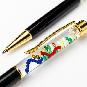 Op de afbeelding: Een zwarte balpen met goudkleurige accenten en een transparant gedeelte in de houder met kleine, kleurrijke drijvende kralen in rood, blauw, groen, wit en goud. De pen is voorzien van een goudkleurige clip en een metalen punt op een effen achtergrond.