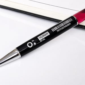 Peut inclure: Un stylo à bille noir avec une pointe argentée et une partie supérieure rouge translucide. Le corps porte l'inscription blanche O+ Rh, HUMAN, 20 mL, ainsi qu'un code-barres avec le numéro 78731301983801 imprimé sur le côté du boîtier noir.