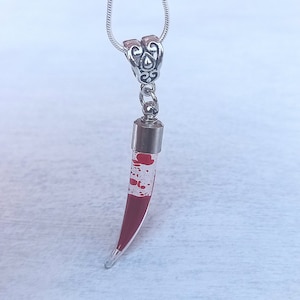 May include: A silver-toned necklace with a pendant shaped like a fang. The pendant is clear with red splatter-like details and a silver cap. The necklace chain is a thin, silver snake chain.
