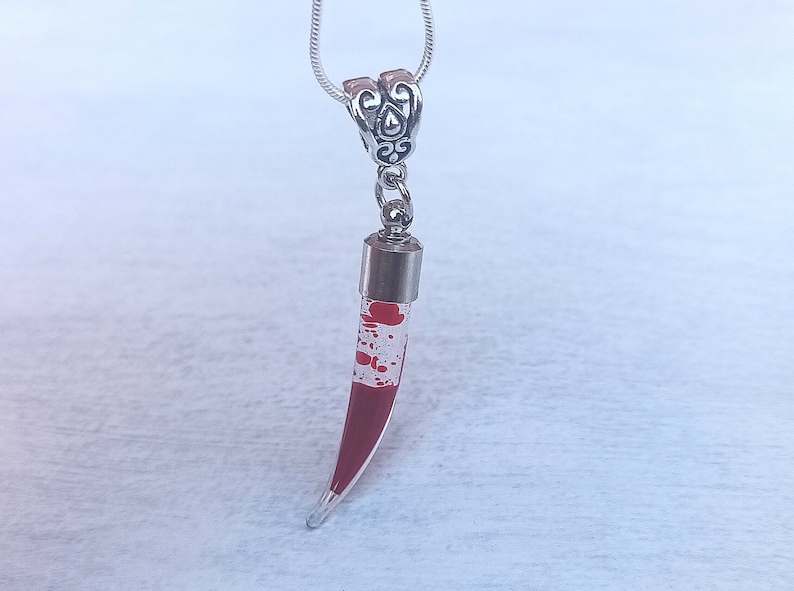 May include: A silver-toned necklace with a pendant shaped like a fang. The pendant is clear with red splatter-like details and a silver cap. The necklace chain is a thin, silver snake chain.