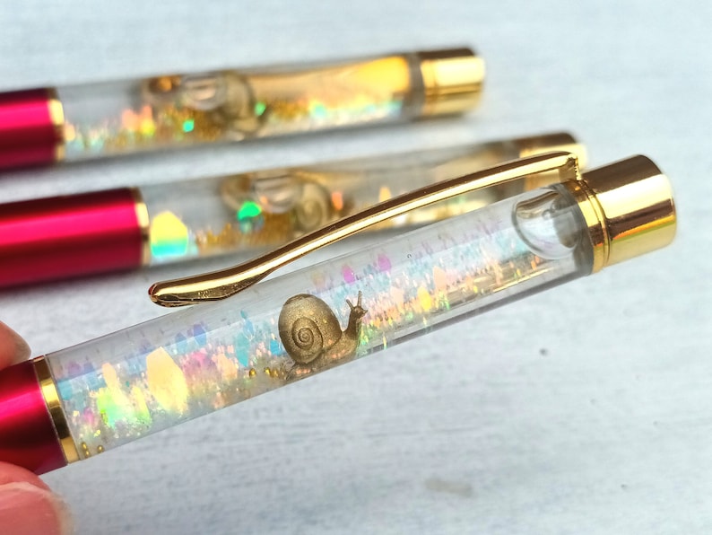 May include: A clear pen with a gold top and a red bottom. The pen contains a gold snail and iridescent glitter.