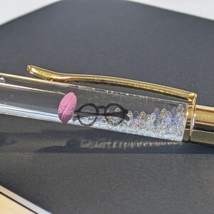 May include: A clear pen with a gold clip and accents. Inside the pen, there is a pink feather, black glasses, and small iridescent beads. The pen is resting on a dark surface.