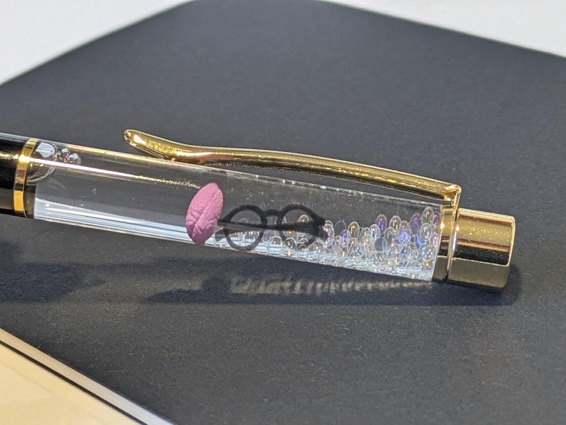 May include: A clear pen with a gold clip and accents. Inside the pen, there is a pink feather, black glasses, and small iridescent beads. The pen is resting on a dark surface.