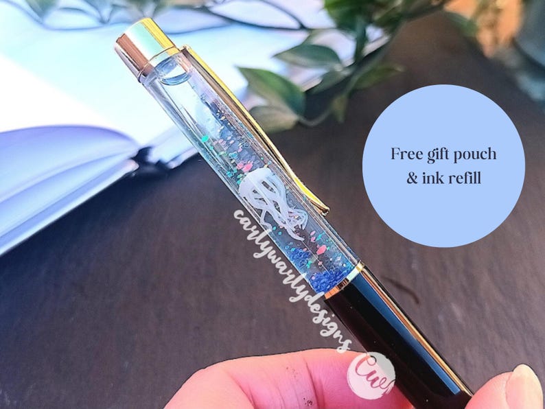 May include: A clear pen with gold accents and a glittery blue and pink liquid inside. The pen has a white design inside the liquid. The text "carlywarlydesigns" is visible on the pen. The text "Free gift pouch & ink refill" is visible in a blue circle.