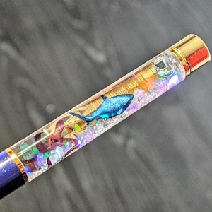 Könnte beinhalten: Ein Stift mit einem transparenten Schaft, gefüllt mit bunten Glitzer, einer blauen Hai-Figur und anderen dekorativen Elementen. Der Stift hat eine goldfarbene Spitze und einen dunkelblauen, glitzernden Sockel.