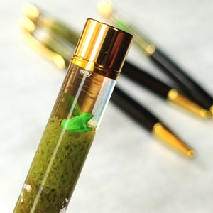 Op de afbeelding: Een transparante pen met een gouden dop en een groene kikker erin. De pen is gevuld met een groene substantie die eruitziet als mos.