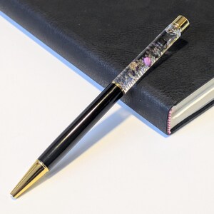 May include: A black and gold pen with a clear barrel filled with decorative elements, including small purple accents. The pen is resting on a black leather-bound notebook with a pink edge. The pen's tip and top are gold.