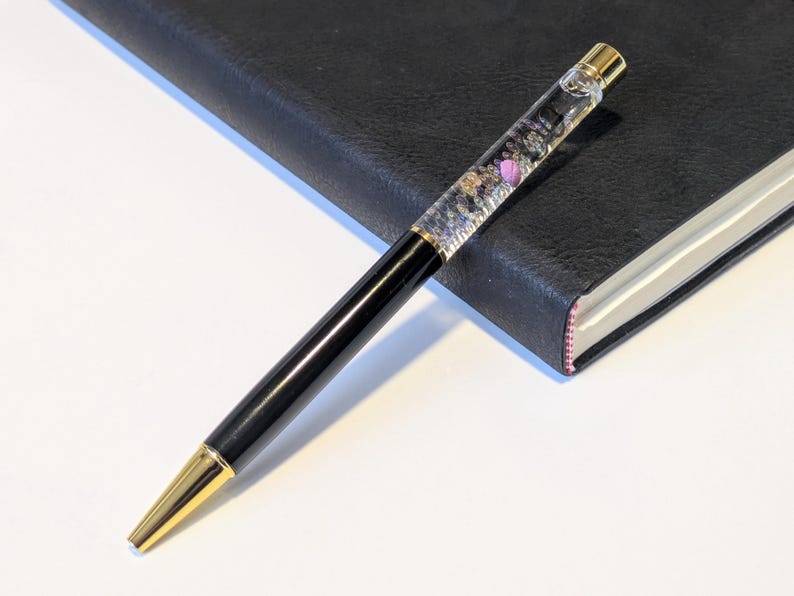 May include: A black and gold pen with a clear barrel filled with decorative elements, including small purple accents. The pen is resting on a black leather-bound notebook with a pink edge. The pen's tip and top are gold.