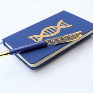 May include: A blue notebook featuring a gold DNA design and a blue ribbon bookmark. A pen with a gold tip and a DNA-themed barrel lies on the notebook. The pen has a gold clip.