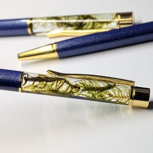 Op de afbeelding: Close-up van drie pennen met blauwe glittervaten en gouden accenten. De doorzichtige penvaten bevatten groen plantmateriaal. De pennen liggen op een witte ondergrond.