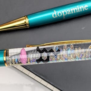 May include: Two teal and gold pens. One pen has the word "dopamine" printed on it. The other pen has a clear barrel filled with small iridescent beads, a pink shape, and a black symbol.