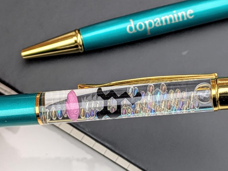 May include: Two teal and gold pens. One pen has the word "dopamine" printed on it. The other pen has a clear barrel filled with small iridescent beads, a pink shape, and a black symbol.