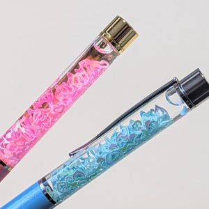 May include: Two pens with clear barrels filled with heart-shaped confetti. One pen is pink with a gold-coloured tip, and the other is blue with a silver-coloured tip. Both pens have a metal clip.