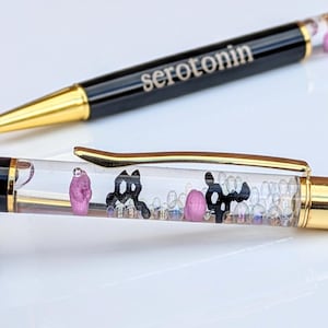 May include: Black and gold ballpoint pens with the word "serotonin" printed on the barrel. The clear barrel contains small decorative elements, including pink and black shapes, and tiny beads. The pen has a gold-colored tip and clip.