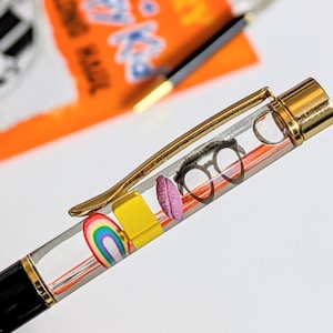 May include: A black and gold pen with a clear barrel filled with colorful miniature objects, including a rainbow, glasses, and a yellow block. The pen has a gold clip and accents.