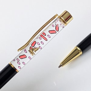 Pode incluir: Duas canetas pretas e douradas. Uma caneta tem um corpo transparente preenchido com formas vermelhas e rosa e um pequeno pingente dourado. A outra caneta tem uma ponta dourada.
