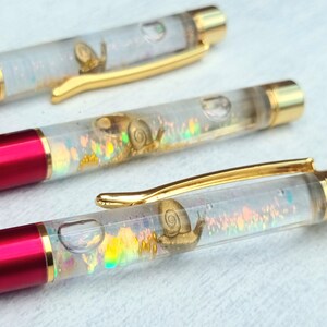 May include: Three red and gold pens with clear barrels filled with iridescent glitter and small snail figurines.