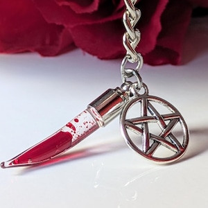 Peut inclure: Un porte-clés avec un petit poignard transparent rempli de liquide rouge et un charme pentagramme argenté. Le poignard a un capuchon argenté et est attaché à une chaîne argentée. Une rose rouge est en arrière-plan.