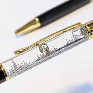 May include: Two black and gold pens. One pen has a clear barrel with a gold clip and a decorative element inside. The other pen is in the background with a gold tip. The pens are on a white surface.