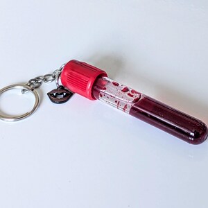 May include: A novelty keychain featuring a clear vial filled with a dark red liquid, resembling blood. The vial has a red cap and is attached to a silver keyring with a small charm of vampire lips. The background is white.
