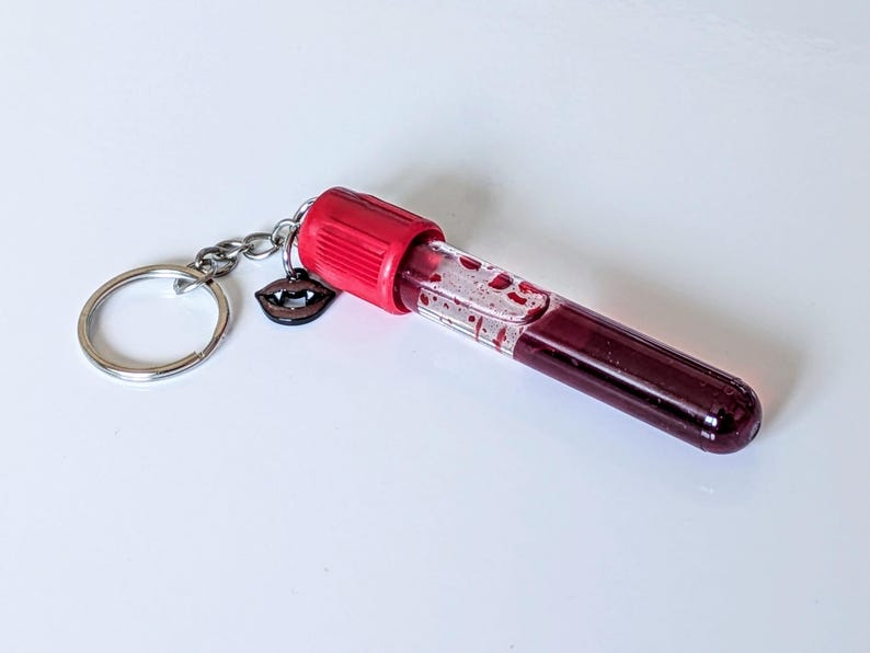 May include: A novelty keychain featuring a clear vial filled with a dark red liquid, resembling blood. The vial has a red cap and is attached to a silver keyring with a small charm of vampire lips. The background is white.