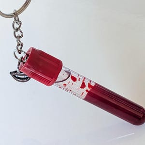 May include: A novelty keychain featuring a clear vial filled with a red liquid, resembling blood, with red accents. The vial is attached to a silver chain and keyring, and includes a small charm of a smiling mouth.