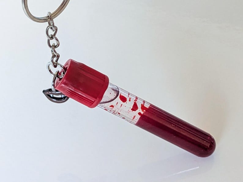 May include: A novelty keychain featuring a clear vial filled with a red liquid, resembling blood, with red accents. The vial is attached to a silver chain and keyring, and includes a small charm of a smiling mouth.