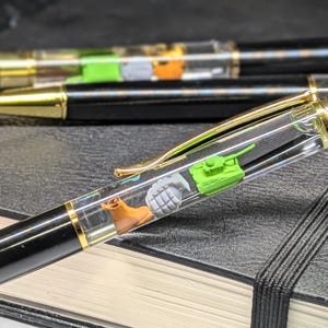 May include: A black and gold pen with a clear barrel filled with miniature figurines. The figurines include a green tank, a brown boot, and a gray hand grenade.