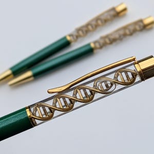 Könnte beinhalten: Zwei grüne und goldene Stifte mit einem DNA-Helix-Design. Die Stifte haben einen transparenten Schaft mit einem goldfarbenen DNA-Modell im Inneren. Die Stiftspitzen und Akzente sind goldfarben. Die Stifte liegen auf einer weißen Oberfläche.