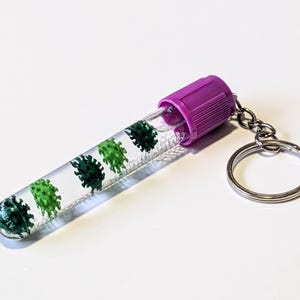 Op de afbeelding: Een novelty sleutelhanger met een doorzichtige buis met groene en donkergroene virusvormige figuren erin. De buis heeft een paarse dop en is bevestigd aan een zilveren sleutelring. De achtergrond is effen wit.