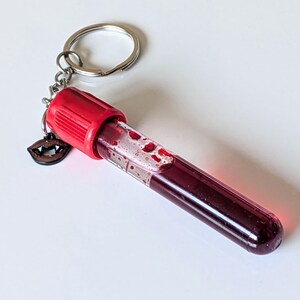 May include: A novelty keychain featuring a clear vial filled with a deep red liquid, resembling blood. The vial has a red cap and is attached to a silver keyring. A small wooden charm of vampire fangs hangs from the keyring.