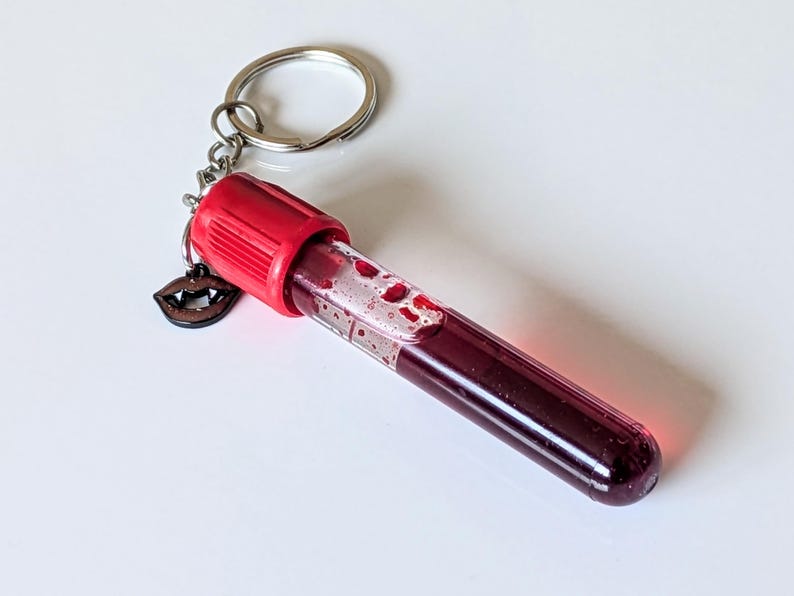 May include: A novelty keychain featuring a clear vial filled with a deep red liquid, resembling blood. The vial has a red cap and is attached to a silver keyring. A small wooden charm of vampire fangs hangs from the keyring.