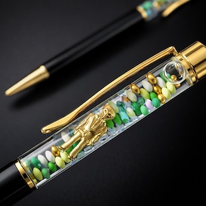 Könnte beinhalten: Zwei elegante Stifte mit schwarzen Schäften und goldenen Akzenten. Ein Stift hat einen transparenten Schaft, der mit bunten Perlen und einer goldenen Figur gefüllt ist. Der andere Stift ist schwarz mit einer goldenen Spitze.