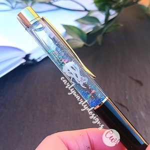 May include: A clear pen with a gold top and black barrel. The pen has glitter and a white design inside the clear section. The text "carlywarlydesigns" is visible on the pen.