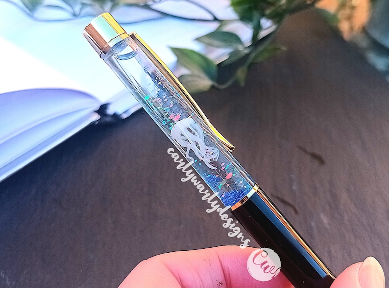 May include: A clear pen with a gold top and black barrel. The pen has glitter and a white design inside the clear section. The text "carlywarlydesigns" is visible on the pen.