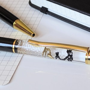 May include: Two pens are shown, one black with gold detailing and the other clear with gold trim, containing decorative elements. A black notebook is in the background, along with a grid-lined paper.