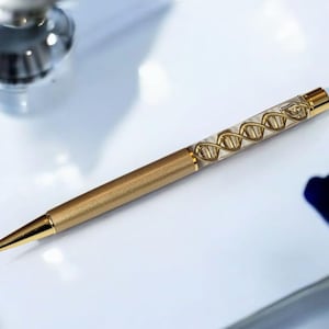 May include: A gold-coloured pen featuring a DNA helix design within a clear barrel. The pen has a sleek, metallic finish and a gold-coloured tip. It is a writing instrument.
