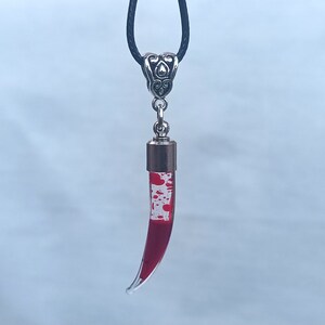 May include: A pendant necklace featuring a clear, horn-shaped glass vial filled with red liquid and white speckles, suspended from a black cord. The vial is capped with a silver-colored metal top and a decorative bail.