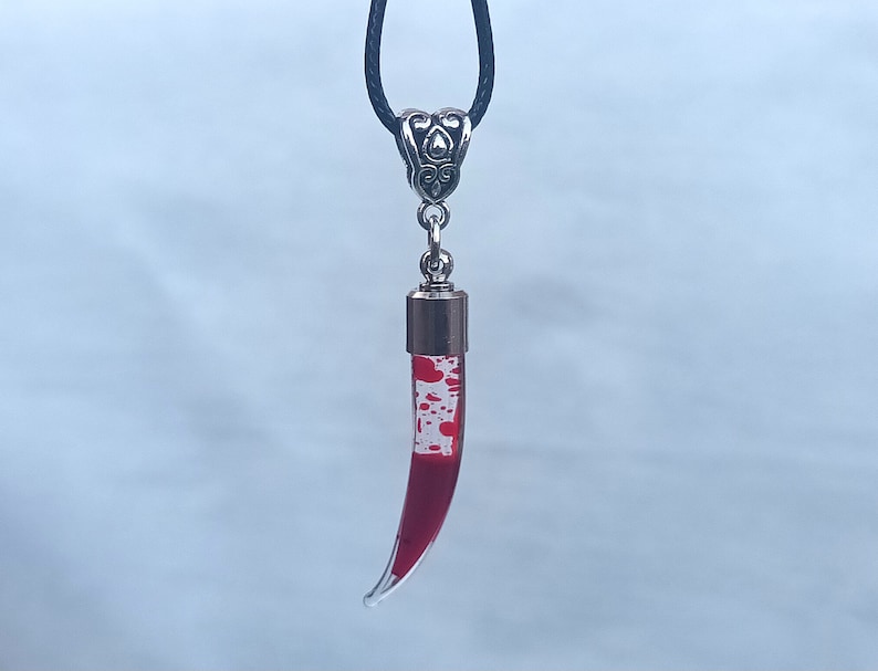 May include: A pendant necklace featuring a clear, horn-shaped glass vial filled with red liquid and white speckles, suspended from a black cord. The vial is capped with a silver-colored metal top and a decorative bail.