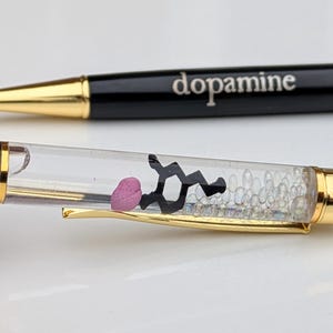 May include: Two black pens with gold accents. One pen has the word "dopamine" printed on it. The other pen has a clear barrel with a pink shape, a black molecular structure, and small clear beads inside.