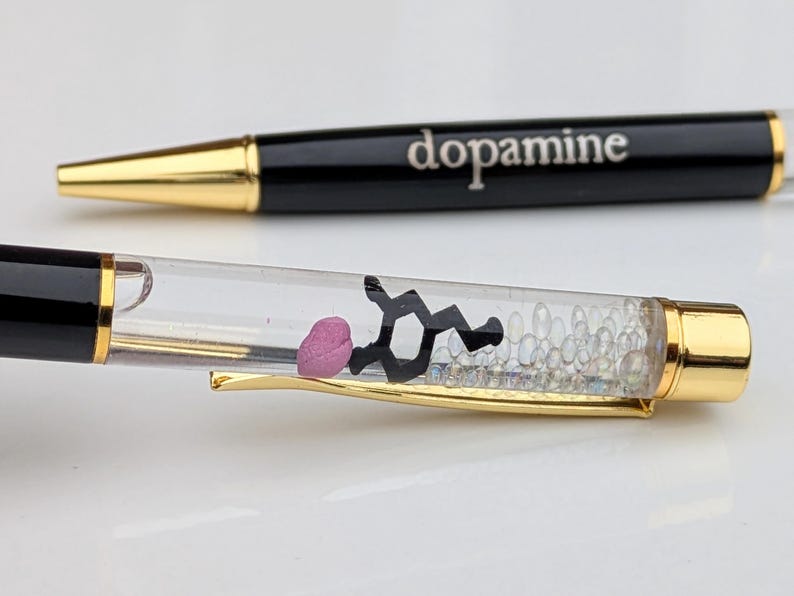 May include: Two black pens with gold accents. One pen has the word "dopamine" printed on it. The other pen has a clear barrel with a pink shape, a black molecular structure, and small clear beads inside.