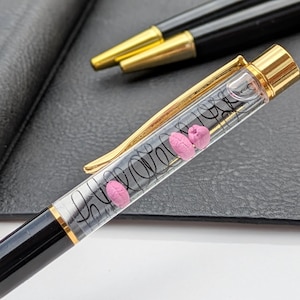 Può includere: Una penna nera e dorata con un fusto trasparente contenente oggetti rosa a forma di cuore e vortici neri. La penna ha una clip e una punta dorate. Un'altra penna è visibile, insieme a un taccuino in pelle nera.