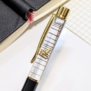 Può includere: Una penna nera e dorata con un fusto trasparente, clip e dettagli dorati. La penna ha un design dorato all'interno del fusto. La penna è appoggiata su una superficie bianca accanto a un taccuino nero.