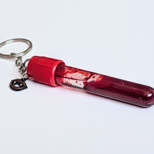 May include: A keychain featuring a clear vial filled with a dark red liquid, possibly a novelty item. The vial has a red cap and a small, dark charm shaped like lips. The keychain ring is silver.