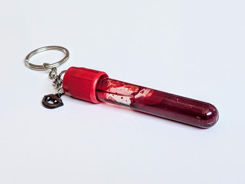 May include: A keychain featuring a clear vial filled with a dark red liquid, possibly a novelty item. The vial has a red cap and a small, dark charm shaped like lips. The keychain ring is silver.