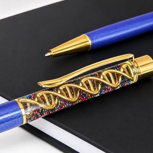 Op de afbeelding: Een balpen met een blauwe metallic behuizing en een transparant bovengedeelte met een goudkleurige DNA-dubbelhelixstructuur omringd door kleine veelkleurige kraaltjes. De pen is voorzien van goudkleurige metalen accenten, een clip en een dop.