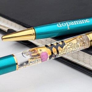 May include: Two teal and gold pens with the word "dopamine" printed on one. One pen has a clear barrel filled with gold liquid, small bubbles, and a pink rose. The pens rest on a black notebook.